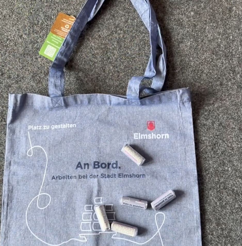 Auf einem grauen Fußboden liegt ein blaugraue Stofftasche mit Werbung der Stadt Elmshorn. Auf dieser liegen kleine Packungen Mentos auf denen Platz zu gestalten steht