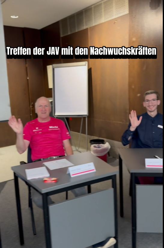 An grauen Sitzungstischen winkt ein Mann mit pinkem Tshirt lächelnd in die Kamera und rechts von ihm winkt ein junger Mann in blauem Hemd lächelnd in die Kamera. Oben steht "Treffen der JAV mit den Nachwuchskräften"