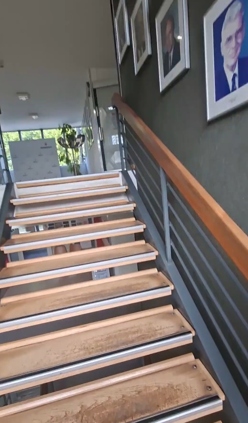 Das Bild zeigt eine Holztreppe in einem Gebäude, rechts an der Wand sind Portraits von Menschen zu erkennen.