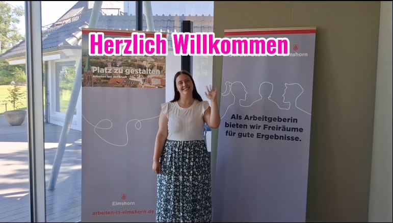 Eine Frau mit langen Haaren, in einem weißen T-Shirt, die bei der Stadt Elmshorn arbeitet winkt.  Herzlich Willkommen steht als Text auf dem Bild