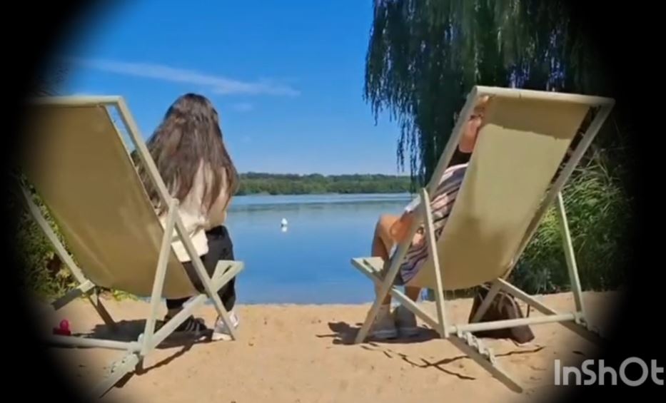 Zwei Liegestühle stehen an einem Strand mit Bilck zu einem See. In den Stühlen sitzen Personen